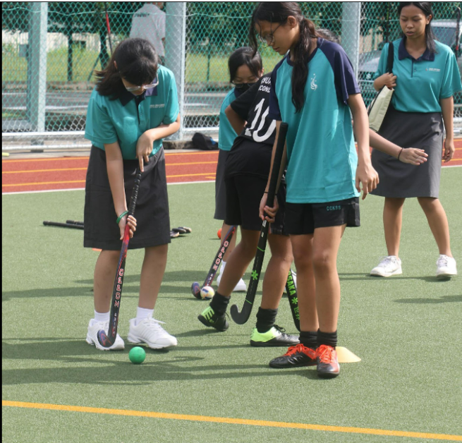 Several girls in teal uniforms holding field hockey sticks stand on a green turf field near a chain-link fence.